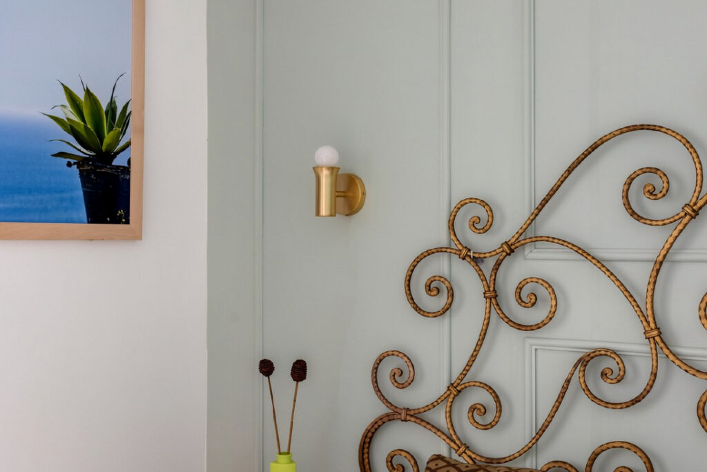 Elegant bedroom detail featuring a curly bronze wrought-iron headboard, a modern brushed-brass wall sconce, and a light sage green paneled wall